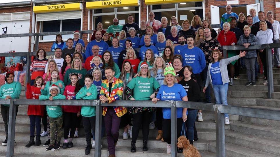 A large group of people, most of them wearing Christmas jumpers, stand on the Blackthorn End terrace at the Memorial Stadium in Bristol. They are smiling and waving at the camera. They are part of Operation Community Hamper which distributes Christmas gifts to families in the Bristol area.