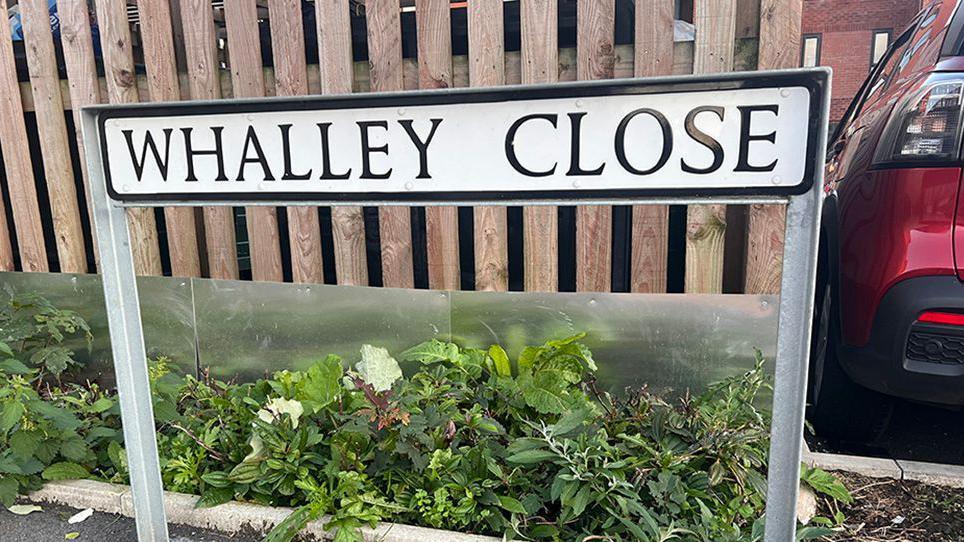 A close-up of Whalley Close street sign with a wooden fence in the background and a dark red car parked to the right.  
