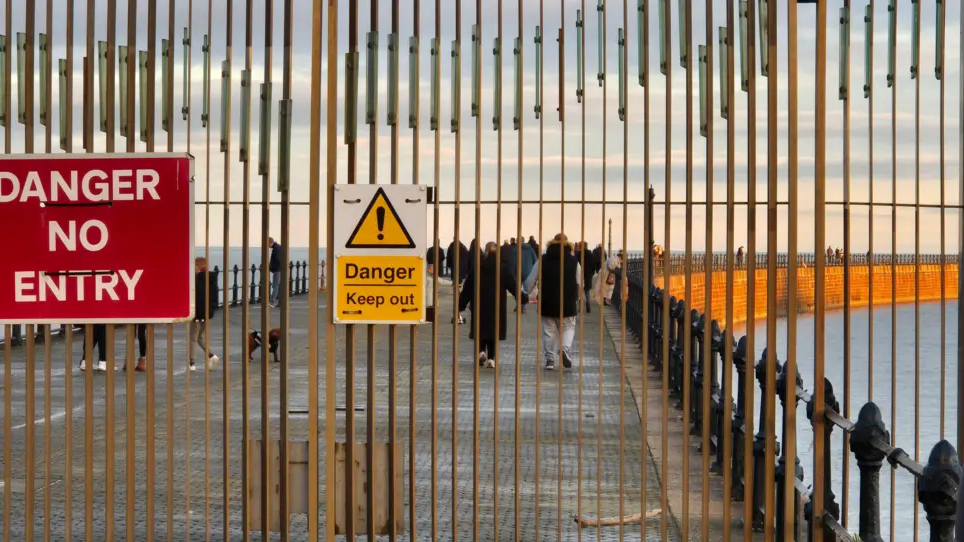 The pier's gates have a red sign reading Danger No Entry and yellow sign reading Danger Keep Out. Dozens of people are strolling along the structure behind the gates as the sun goes down. The sea is calm.