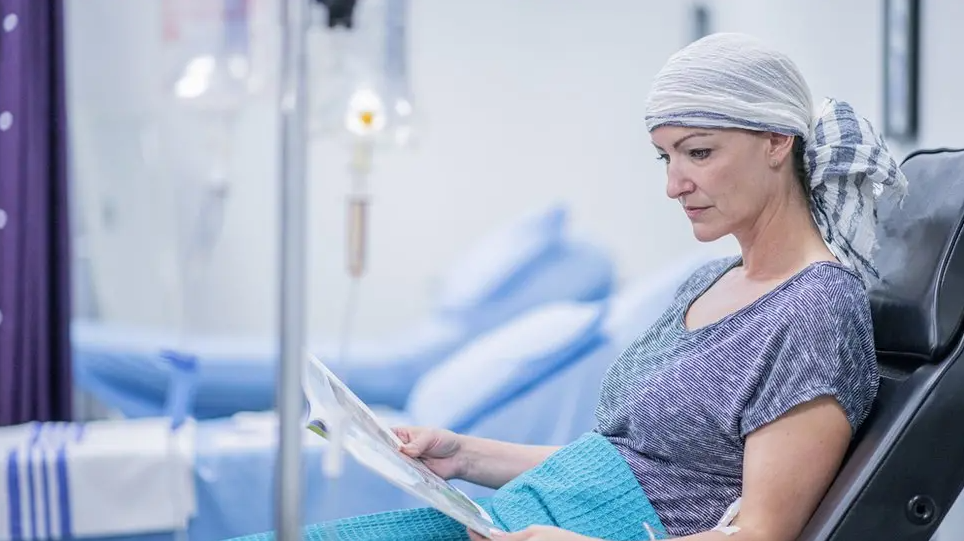 A woman in hospital receiving cancer treatment. She has a head scarf on and is in bed reading.