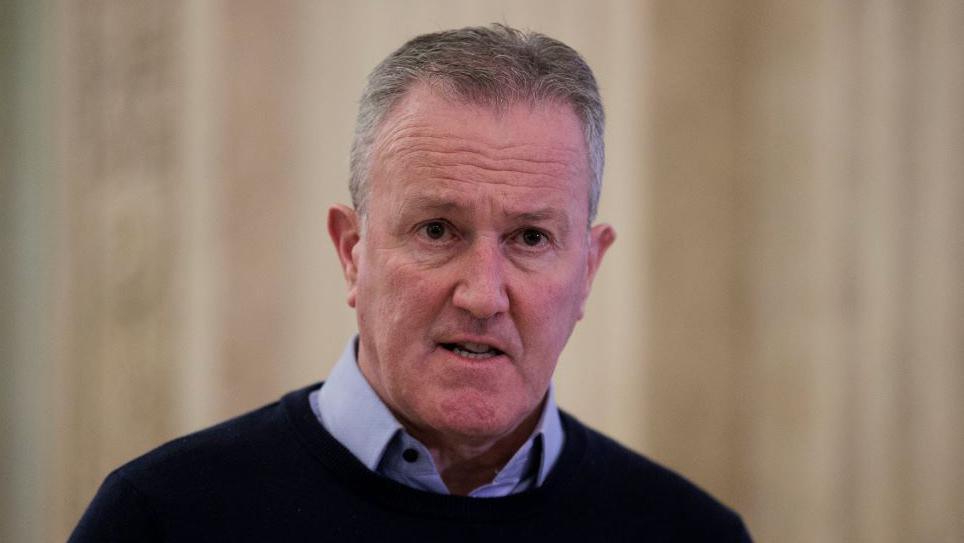 Sinn Fein MLA Conor Murphy stands in Stormont. The background is a blurred beige wall. Murphy is wearing a blue jumper and a blue shirt with the top button undone. 