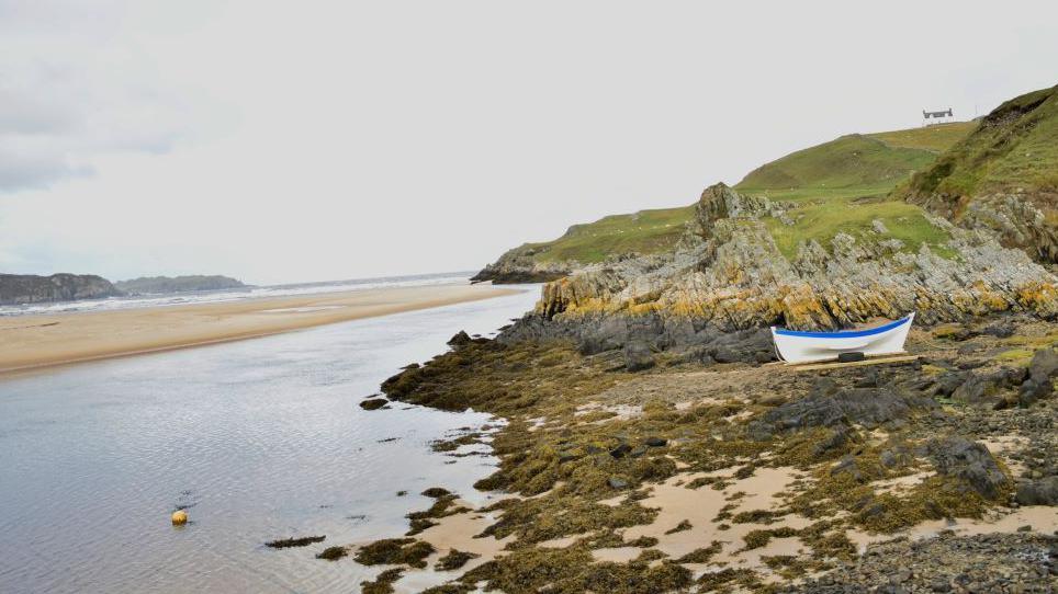 The boat, which is painted blue and white, on a rocky beach near the sea.