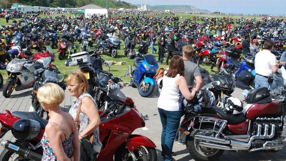 A sea of colourful motorbikes parked on a grassy patch in Ramsey with men and women standing next to them.