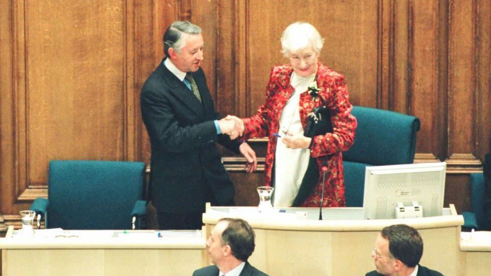 Winnie Ewing shakes hands with Sir David Steel as she hands over the chair to the newly elected presiding officer. She is standing to his right on a raised area which has seats and a computer screen. Two men in front of them have turned to look.