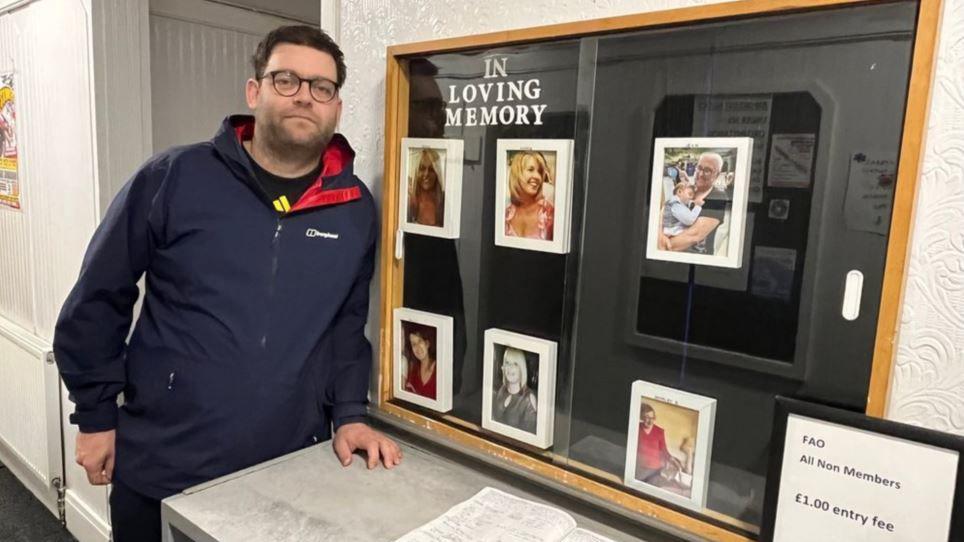 Vice president Tom Steer with short brown hair and glasses in a Berghaus jumper at the signing in desk at the club with photos of people linked to the club who have died 