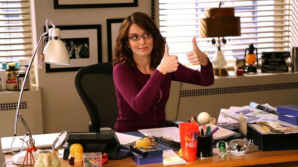 Tina Fey as Liz Lemon in 30 Rock - wearing a stripy sweater ad glasses she sits at her desk on the show, giving a two-thumbs-up sign to someone off to the left.