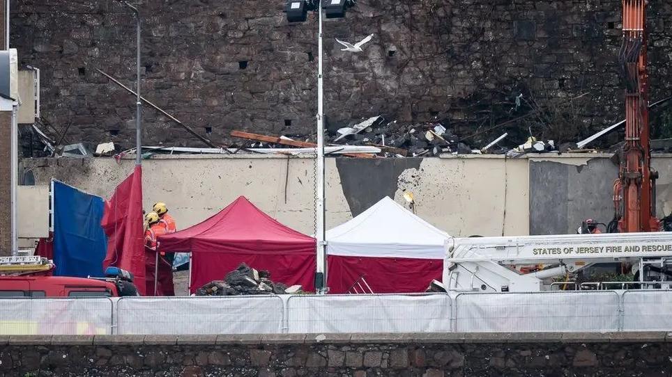 Rubble and aftermath of the fire next to pop-up tents for emergency workers, two of which, wearing safety helmets, are talking with each other 