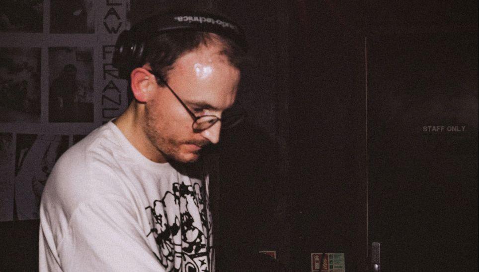 Leigh Dennis is standing in a dark room wearing a white T-shirt and black headphones. He is looking down onto a desk.