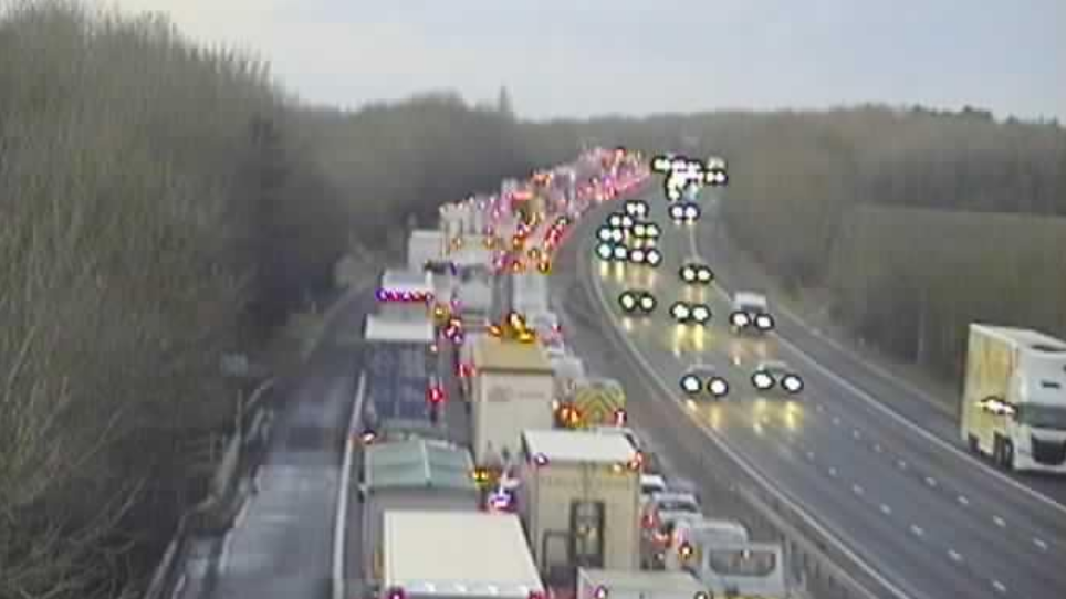 Three lanes of traffic on the M11 southbound can be seen from a cctv motorway camera. The M11 northbound, to the right of where the traffic is, is clear with cars moving easily past. 