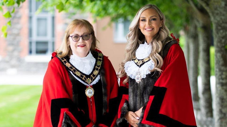 Deputy Mayor Julie Gilmour and Mayor Leah Kirkpatrick pose together wearing red ceremonial robes and their gold chains of office. Kirkpatrick has long, wavy blonde hair.  Gilmour has shoulder-length blonde hair with a fringe and is wearing dark glasses. Both women are smiling, there are trees and a lawn in the background. 