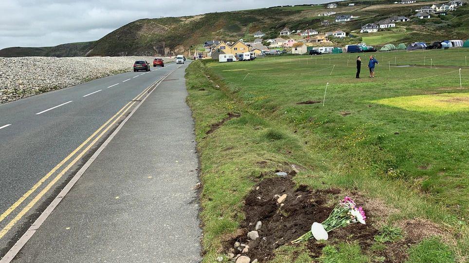 A coastal road right next to fields with camping pitches seen on the grass and one bouquet of flowers. There are houses scattered up the hills on the right side of the image.