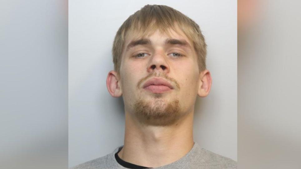 A police custody photo of James Willett. He has short blonde hair with a fringe. He is wearing a black top underneath a grey jumper.