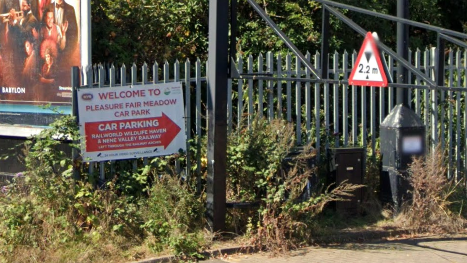 A Google Street View image showing a sign for Pleasure Fair Meadow car park attached to green fencing which is surrounded by overgrown weeds and plantation