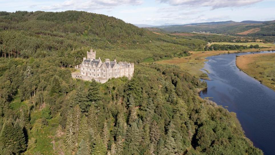 Aerial image of Carbisdale Castle on a tree-covered cliff overlooking a loch