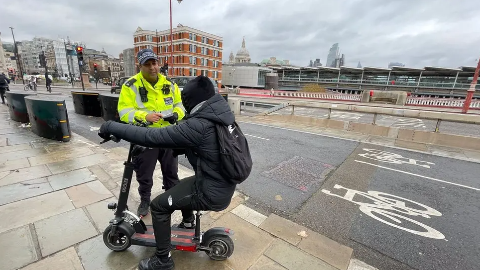 A male with his back to the camera is sat on an e-scooter talking to a police officer in London. The man has dark clothing on and hood. He is on a wet pavement by a cycle lane.