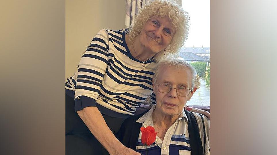 Margaret Tyman with white curly hair wearing a navy, white and beige striped top and navy pants bends down to take a red rose from her husband Donald Tyman who is sitting down with white hair, brown eyes, wearing a navy blue, pale blue and white striped top and beige cardigan