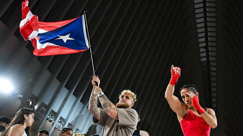 Jake Paul waves a Puerto Rico flag and stands next to Amanda Serrano who is throwing a punch
