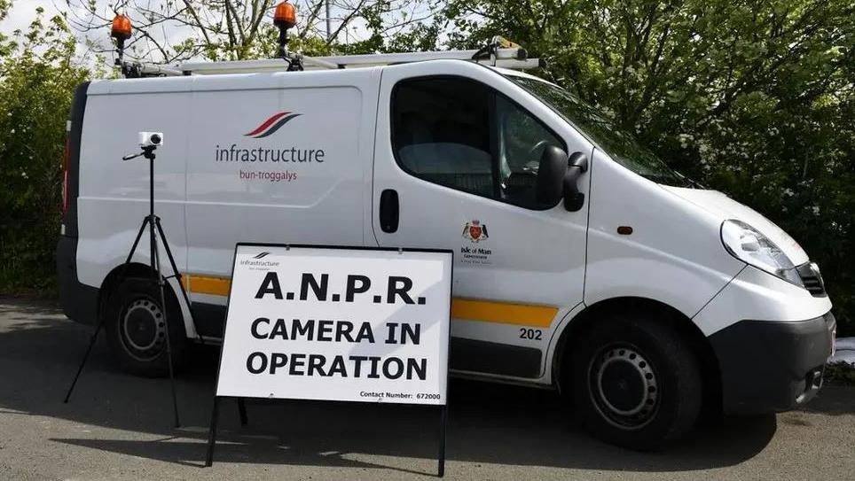 A white van parked on a road near some green trees. It has a DOI logo and a yellow stripe on the side and there is a camera on a tripod in front of it. Next to it there is a white sign with black writing that reads ANPR camera in operation.