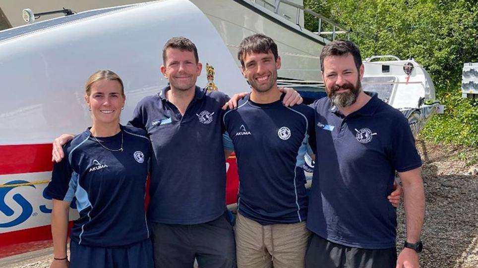 Jess Hunter, Dec Lynn, Gian-Luc Angiolini and Graham Doyle standing smiling in front of their boat. They are all wearing navy casual tops and have their arms round each other