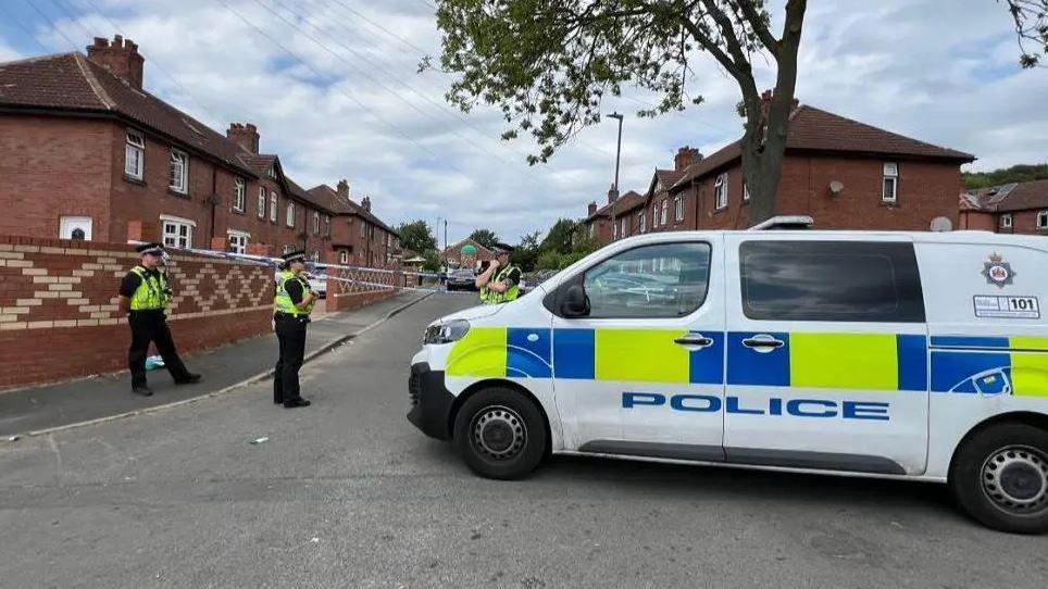 A police van in a street which has been cordoned off. There are three police officers standing nearby.