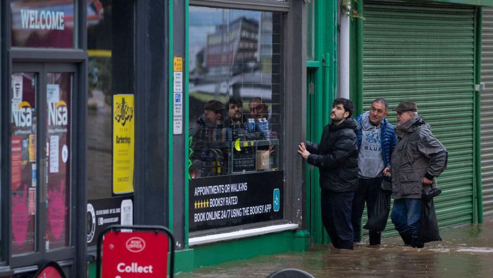 Flooding on Mill Street in Pontypridd. the water is up to the bottom of a shop window at the Nisa store. Three men are standing outside the shop wiht the water up to their knees. 