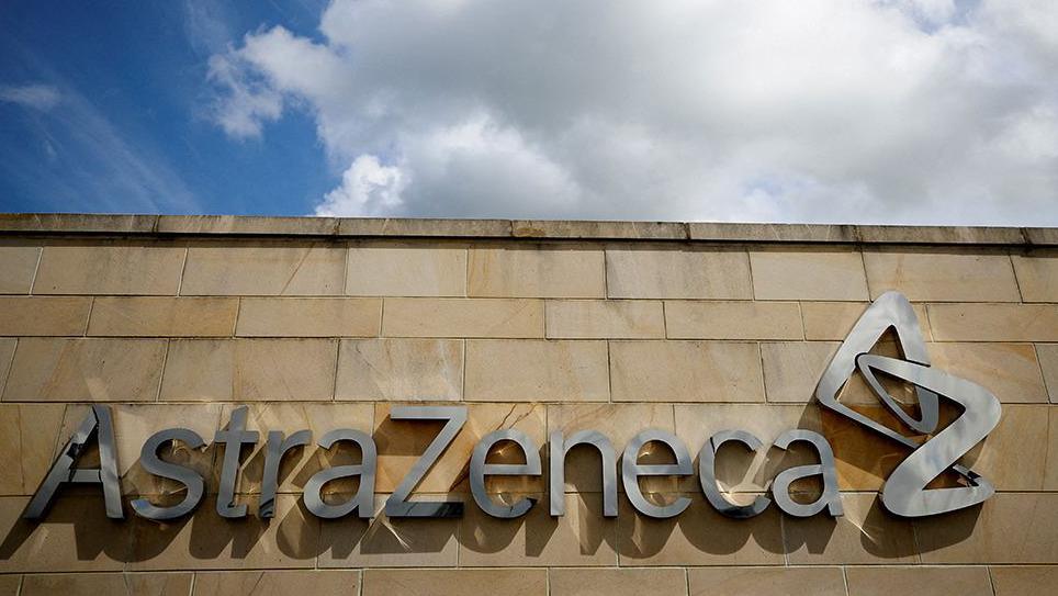 A sign on a building in silver saying AstraZeneca. It is on a sandstone brick wall and above it there is a blue sky with white clouds.