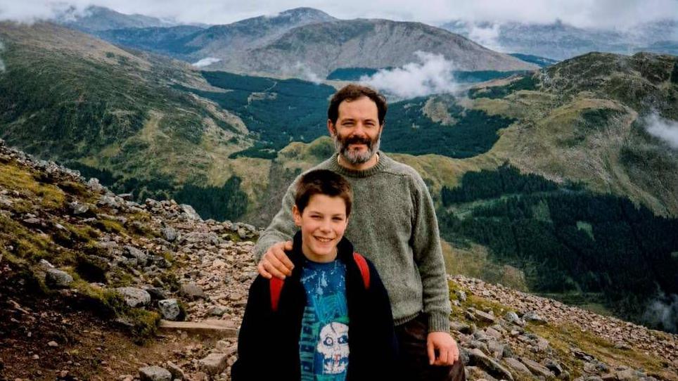 A young boy standing on top of a hill with a blue t-shirt on. The straps of a red backpack are visible over his shoulders. A man with a thick beard has his arm round the boy. They are both smiling.