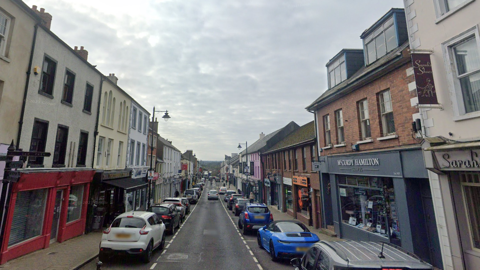 A street view image of Ballymoney town, there are numerous cars parked on either side of the street. There are a range of different shops painted in grey, orange, red and black.