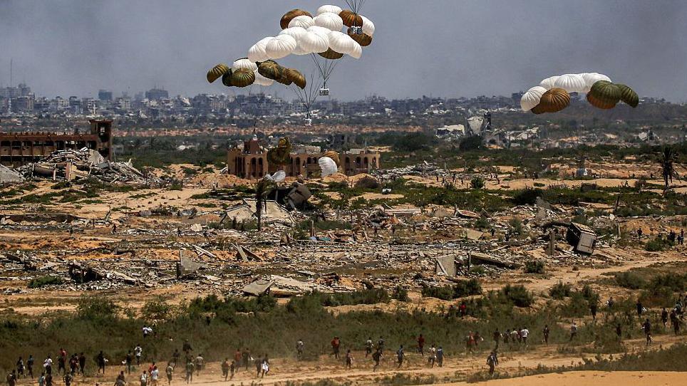 Palestinians rush to the scene as aid pallets are parachuted after being dropped from a military plane over Nuseirat in the central Gaza Strip during an airdrop mission on August 5, 2025. There are several people running towards the aid packages in the foreground and flattened buildings below the falling aid.
