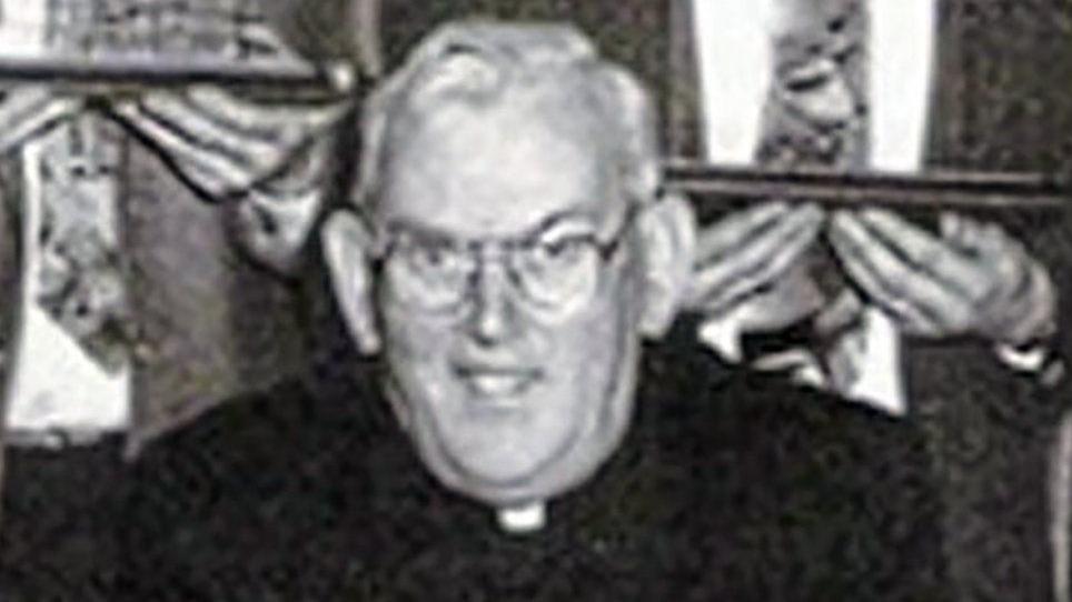 A black and white photo of the late Fr Malachy Finegan. The priest had short white hair and glasses and wore a black suit with a white clerical collar. 