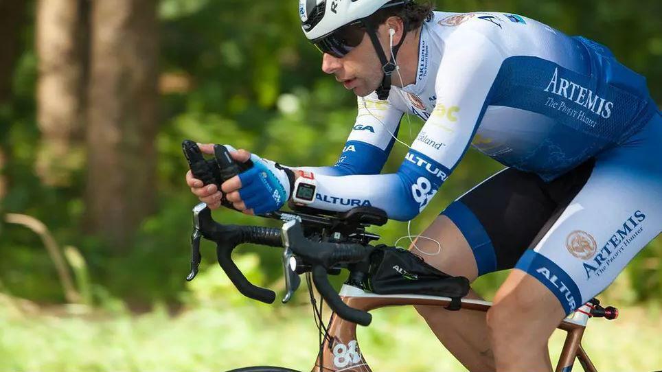 A man in a blue and white lycra cycling outfit rides a bike through a woodland.
