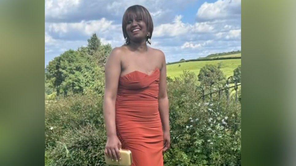 A woman in a red dress is standing outside with hills in the background. She is laughing and holding a small purse.