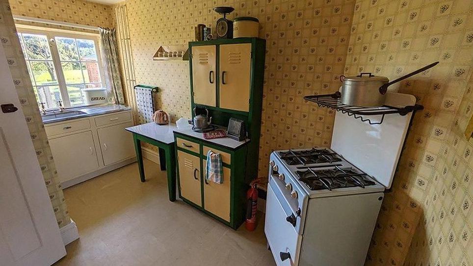 A kitchen from the 1950s with a stand-alone cooker, a dresser and a table. The wallpaper is a pattern of yellow or beige squares.