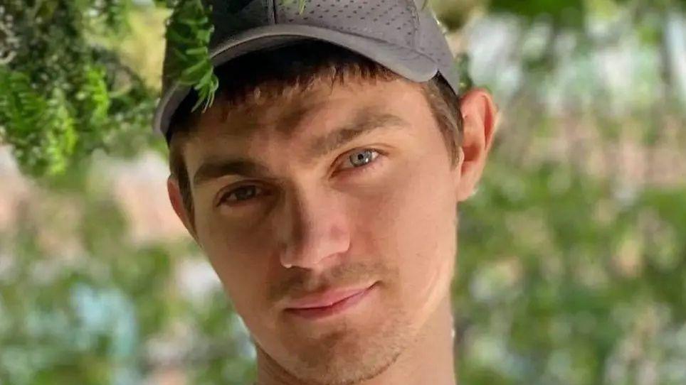 A young man with short brown hair, wearing a grey baseball cap, in pictured outside in some gardens.