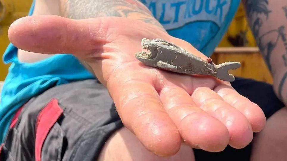 A close up of a person, the shoulders and knees are cropped. The person is wearing a blue vest top and grey shorts with red pockets. A hand is stretched out towards the camera holding a lego shark. 