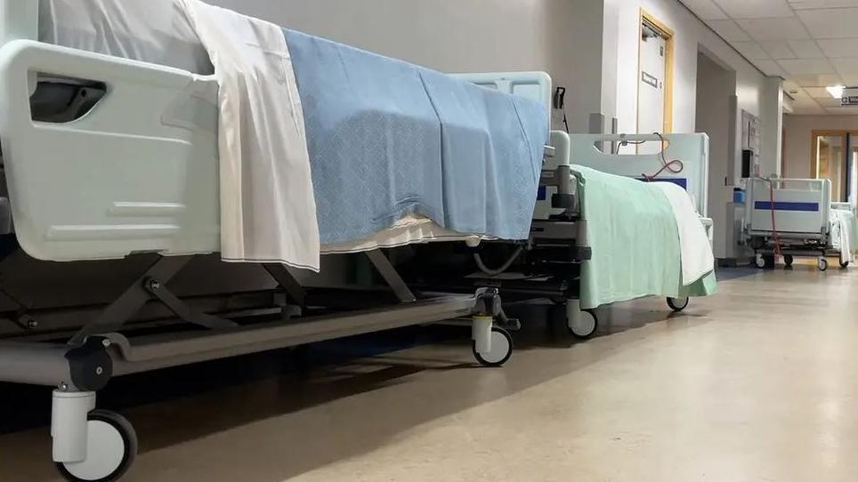 Empty hospital beds line the corridor of a hospital. They are on wheels and have blue and green blankets draped across the top. 