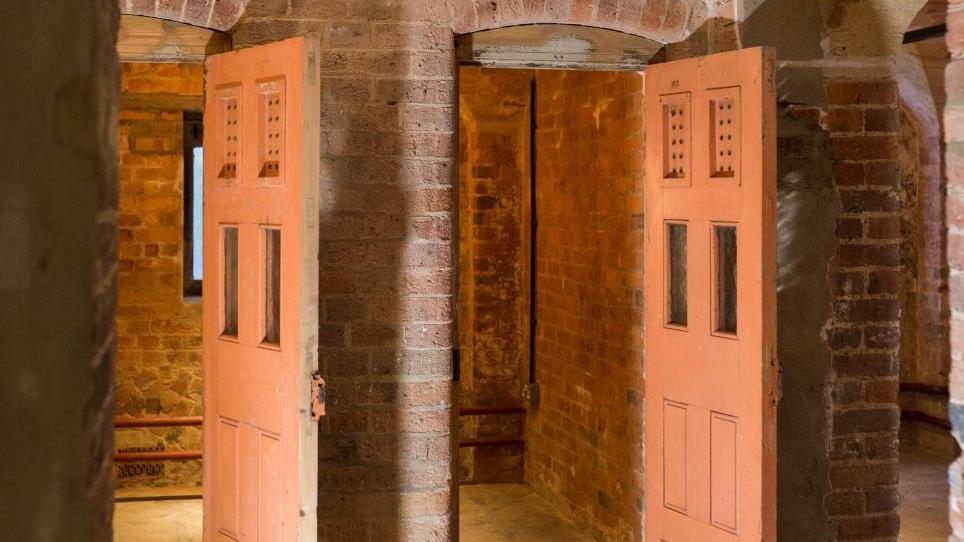 Doors open in subterranean cells at the St Albans Museum and Gallery, which is a former court. The cells are all exposed brick with heavy doogrs with two small panel windows. 