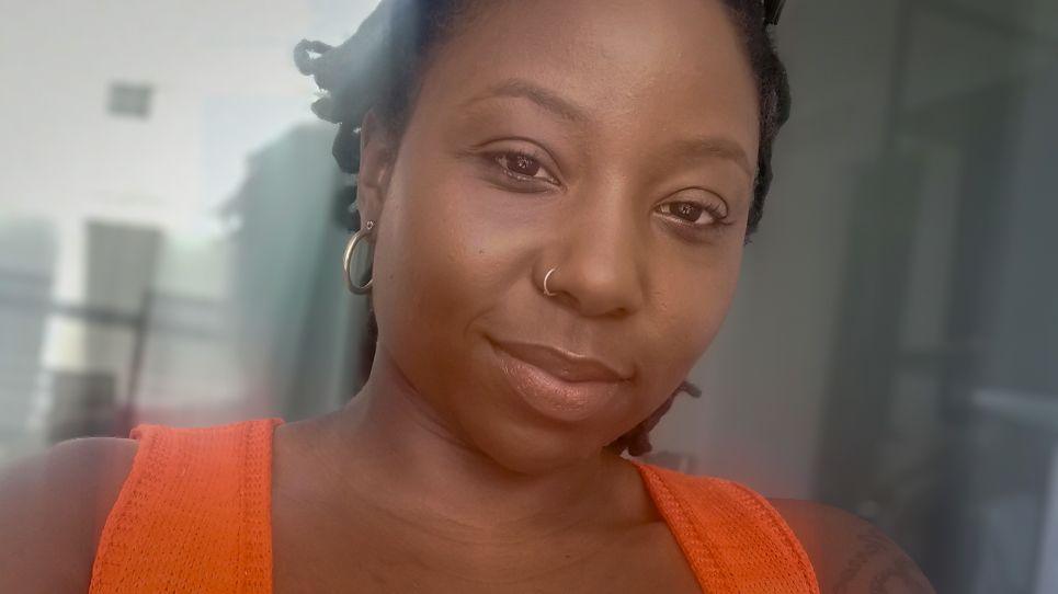 A woman with dark hair and a nose ring, wearing an orange top, smiles at the camera in a selfie