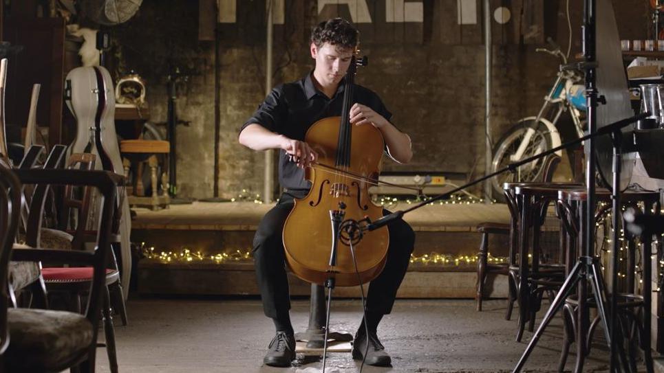 Sam Hills-Field playing a cello. He is wearing black with a stage behind him that has twinkling lights on it and the room is filled with wooden chairs.