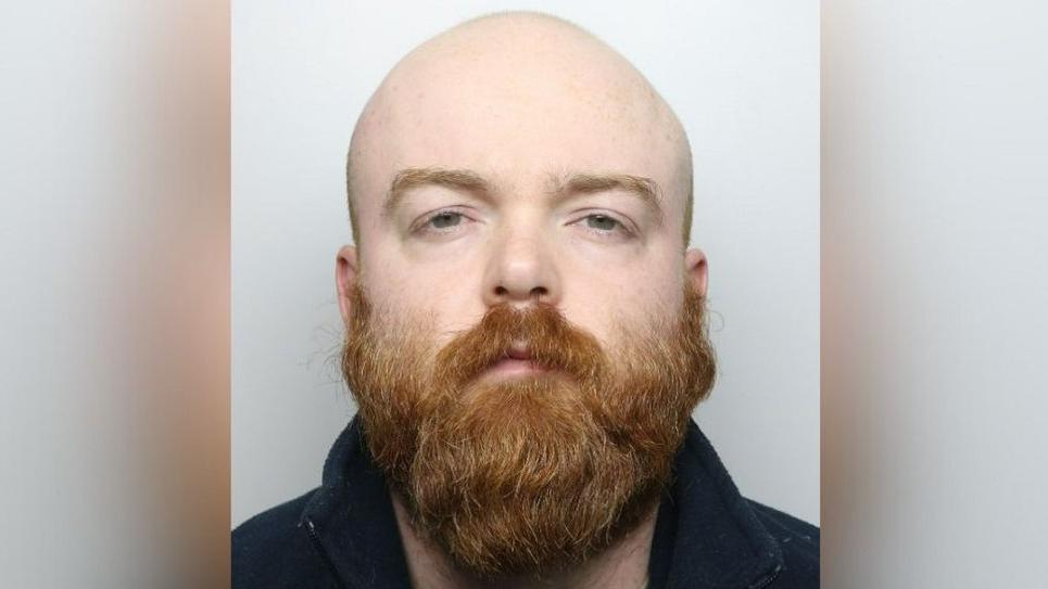 Benjamin Pinchin with a bald head and thick ginger beard. He is wearing a black top and has a stern expression as he stands in front of a grey background. 