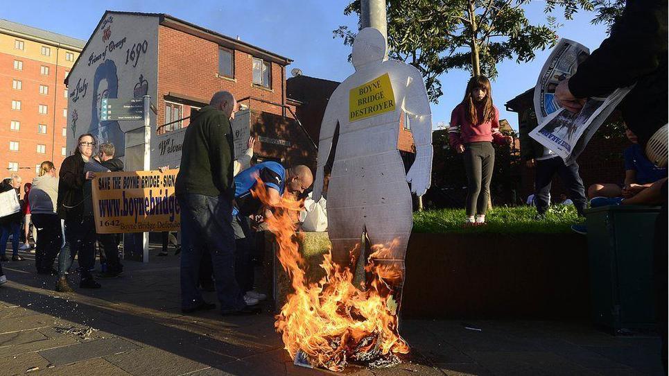 A cardboard cut out in the shape of a man is burned. It says Boyne Bride Destroyer. There are protesters standing around it. A mural of King Billy is to the left.