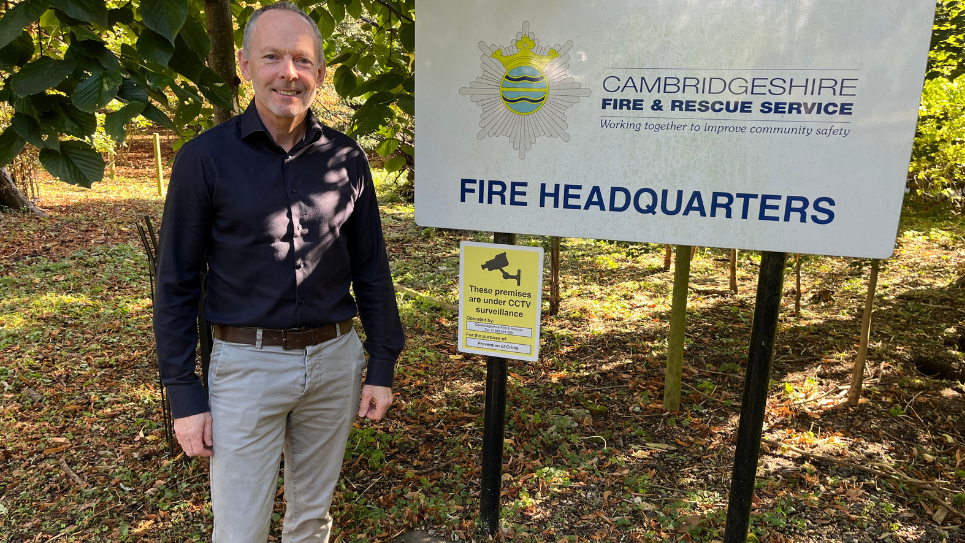 Chris Strickland smiles at the camera dressed in a dark shirt and chinos. He is standing outside next to a sign reading "Cambridgeshire Fire & Rescue Service". 