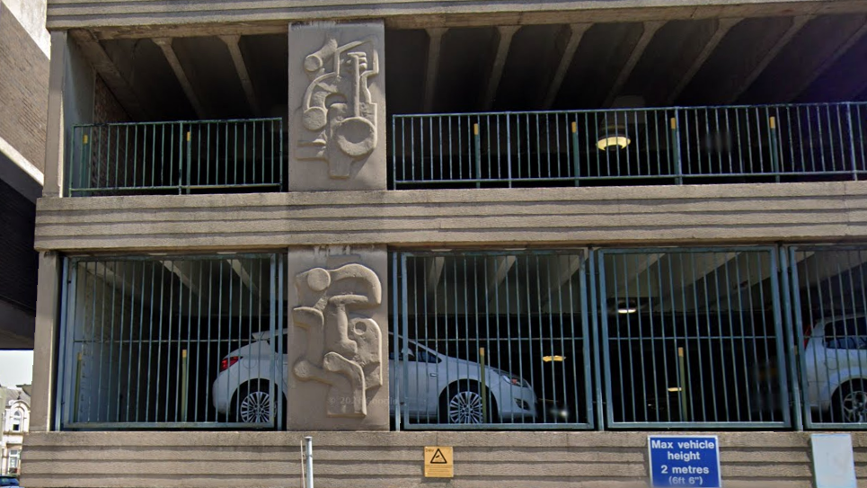 Exterior shot showing two floors of a concrete multi-storey car park showing abstract designs in relief on the pillars and blue metal railings in between. Two cars are visible parked inside on the lower floor.