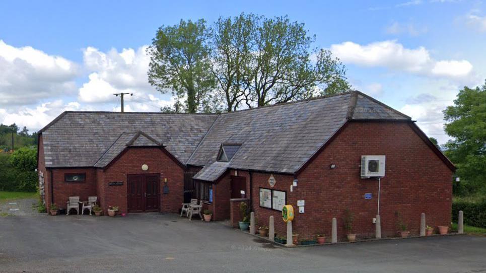 An L-shaped red brick building with a large dark roof, surrounded by bollards and a car park.