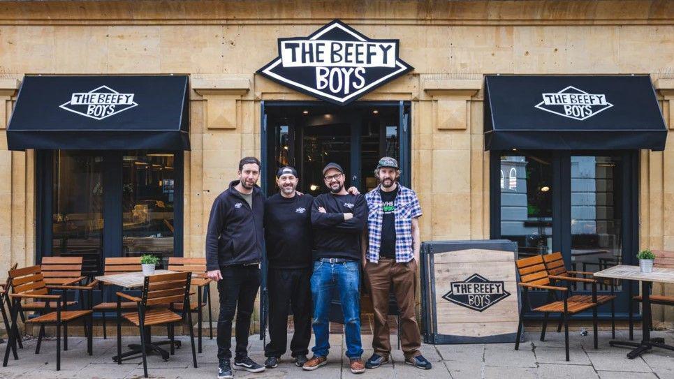 Four men standing outside a storefront built in honey-coloured stone. They are all wearing caps and three have branded restaurant tops on. The fourth is in a checked shirt. Next to them, a sign reads "The Beefy Boys"