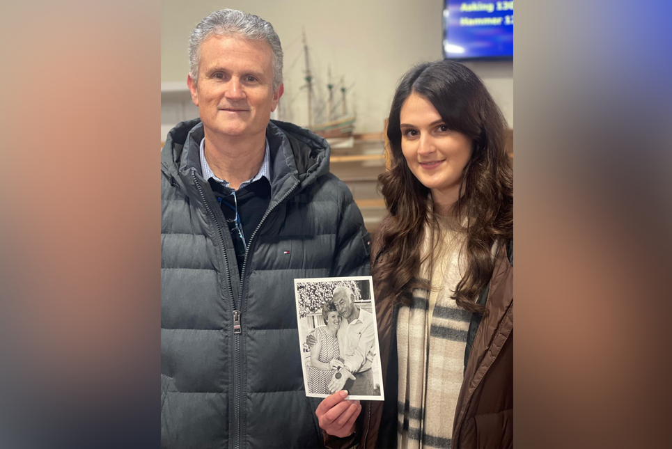 Richard and Hollie Higgin stand in the auction room holding up a black-and-white photo of their grandfather/great-grandfather. Richard has short grey hair and is wearing a blue coat. Hollie has long brown hair and is wearing a brown coat.