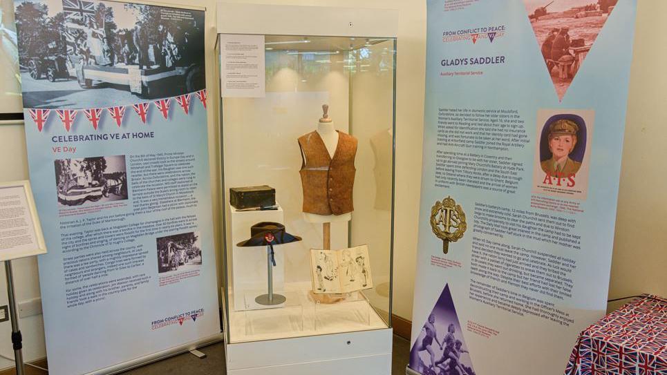 A view of the Soldiers of Oxfordshire Museum exhibition on VE and VJ Day. It hs banners with information on Celebrating VE at Home in 1945 and Gladys Saddler. Several items such as a hat, a vest and a box are behind glass. An edge of a table covered with a union jack cover is beside them.