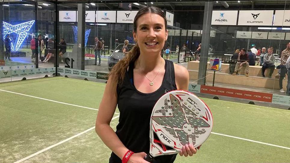 A smiling woman in sports gear standing on a court holding a padel raquet which has holes across it.