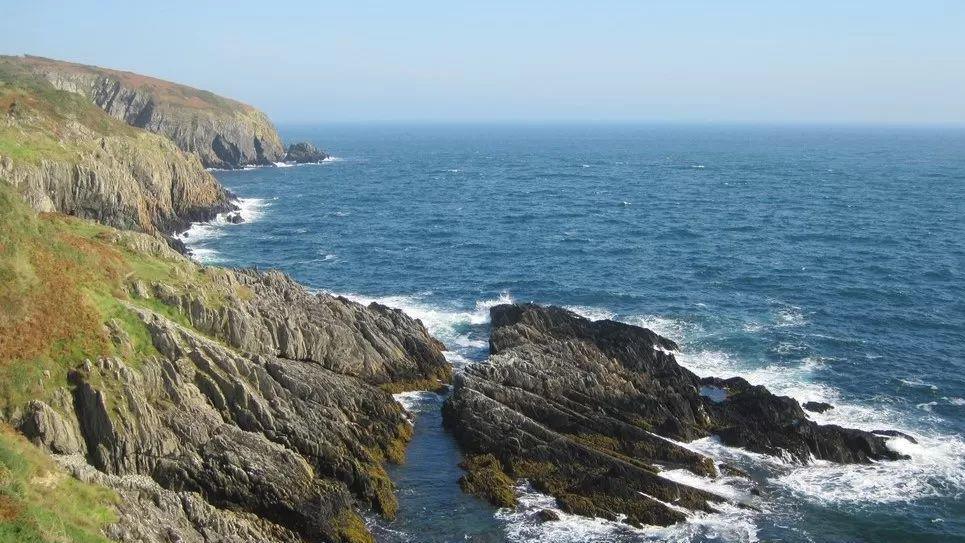 The rugged coastline of the Isle of Man, brown cliffs and rocks with the sea breaking against them..