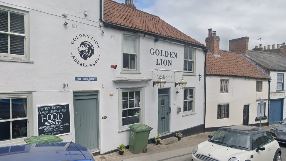 A pub with a white front and blue doors. It has a lion painted above one of the doors. To the left is a blackboard that reads "food served". There are cars parked outside.